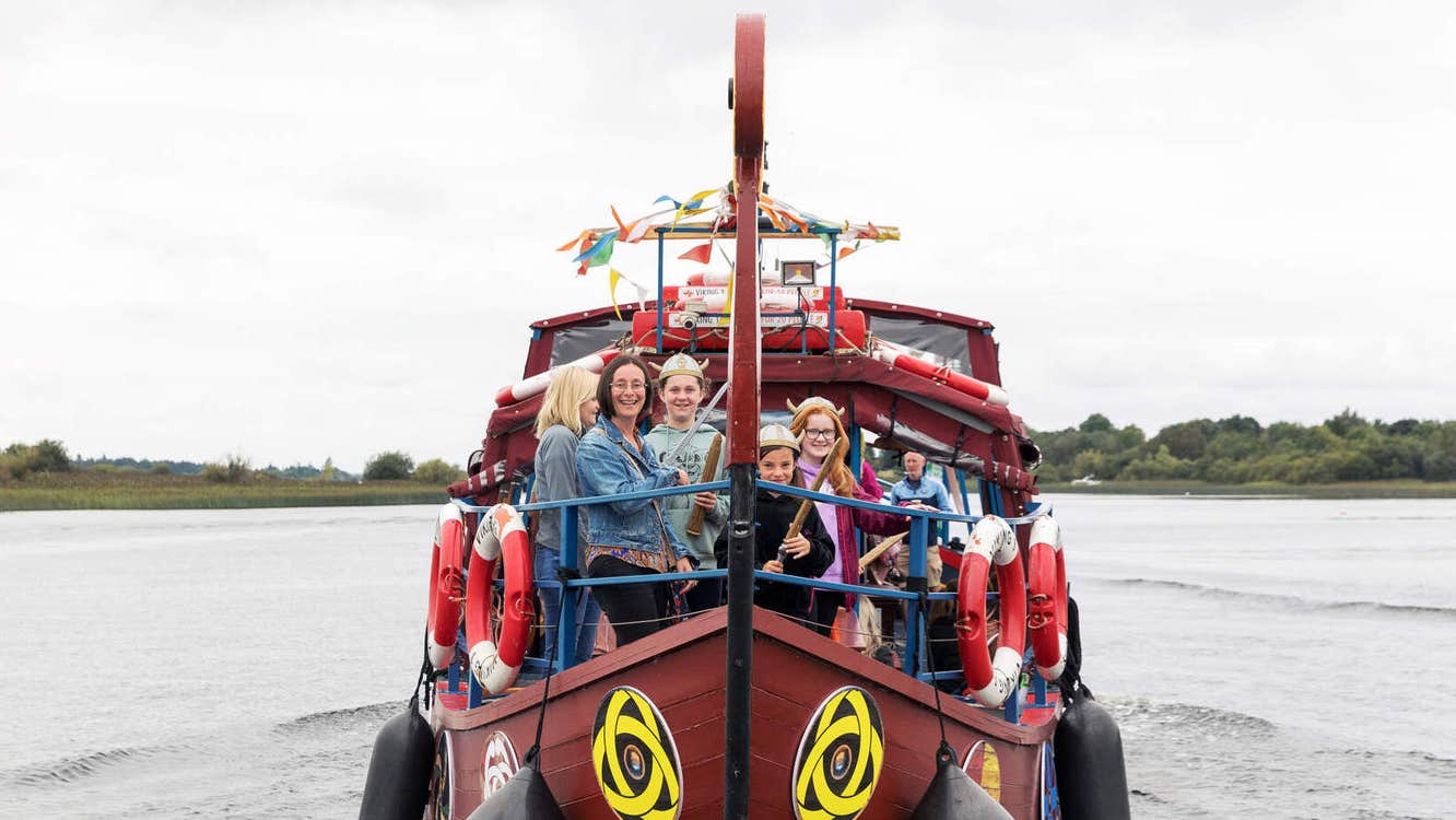 Image of Viking Ship Athlone with people standing at the bow of a small brown ship on the water.