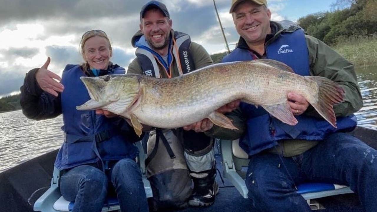 Three people in blue life vests sitting on a boat holding a big fish