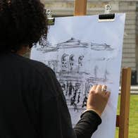 Rear view of person drawing on easel outdoors in front of a building
