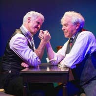2 men in shirts and waistcoats arm wrestling on a small table.