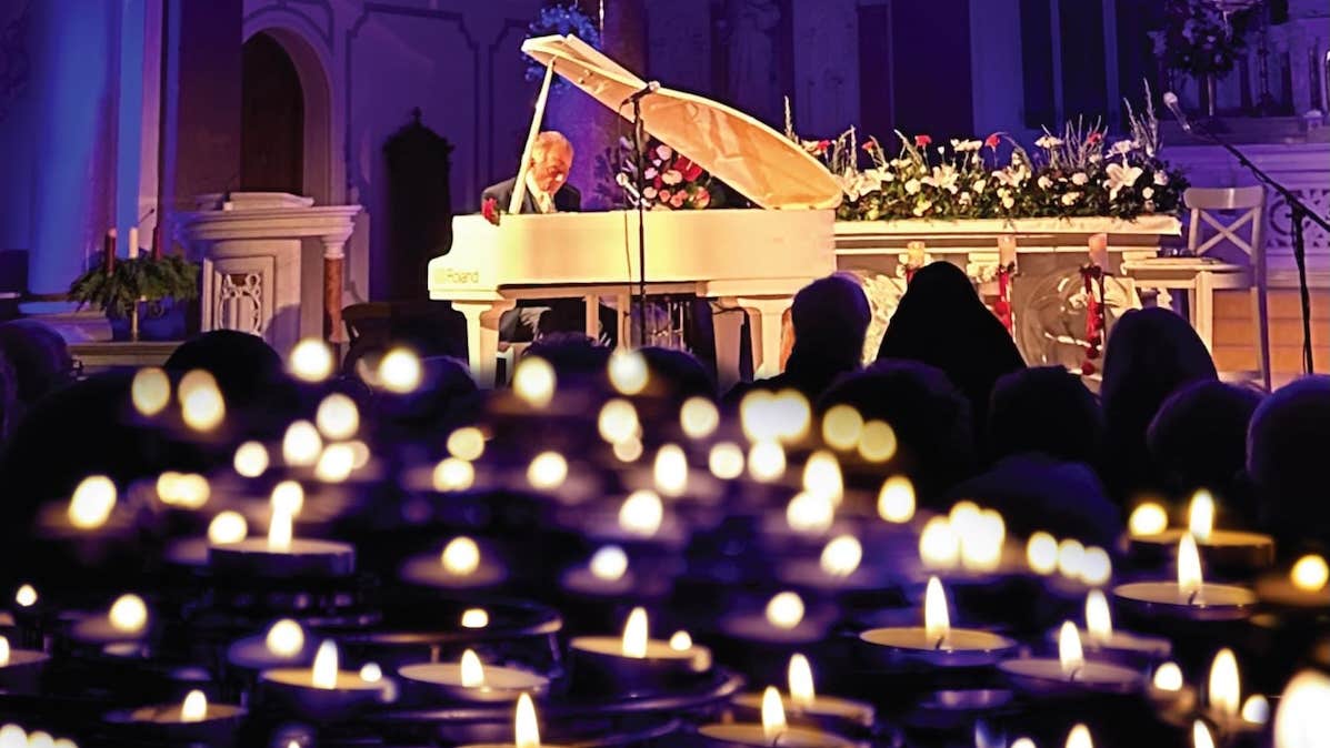 In the distance a man playing a white grand piano with lots of blurry candles in the foreground.