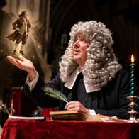 A man in white curly wig is seated at a table with a candle and a tiny figure of a man on his outstretched hand.