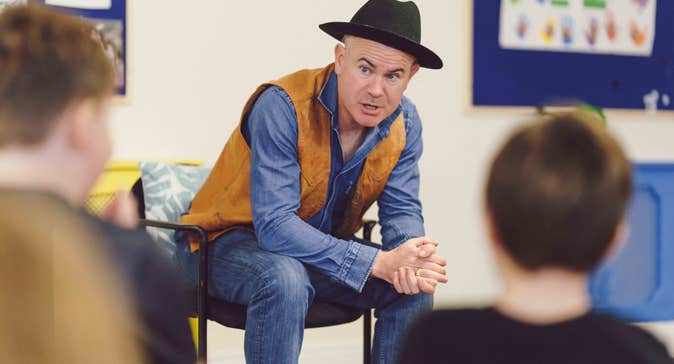 Killian Storytelling for Children - a man in a black hat, seated leaning forward talking, blurred rear view of children in foreground.