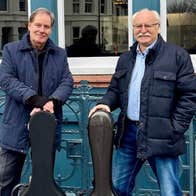 2 older smiling men with guitar cases standing in front of railings.