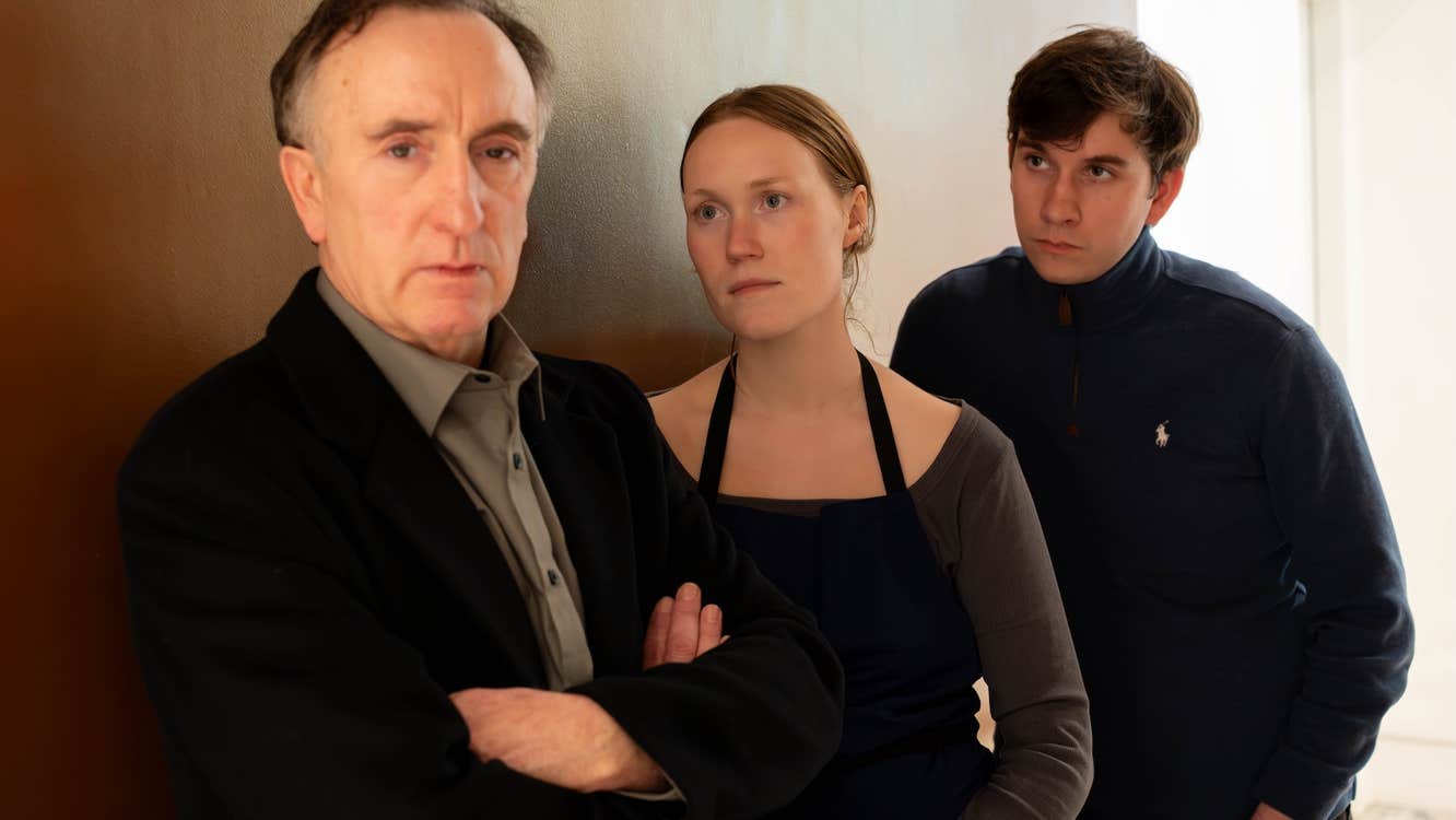 2 people are leaning against an internal wall looking at a man with his arms crossed, not looking at them.