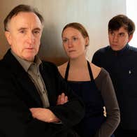 2 people are leaning against an internal wall looking at a man with his arms crossed, not looking at them.