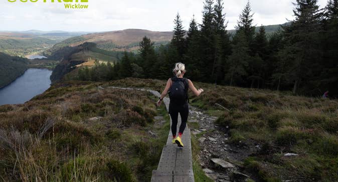 EcoTrail Wicklow 2026 - a person running along a narrow wooden path on rough land with trees and hills in background.