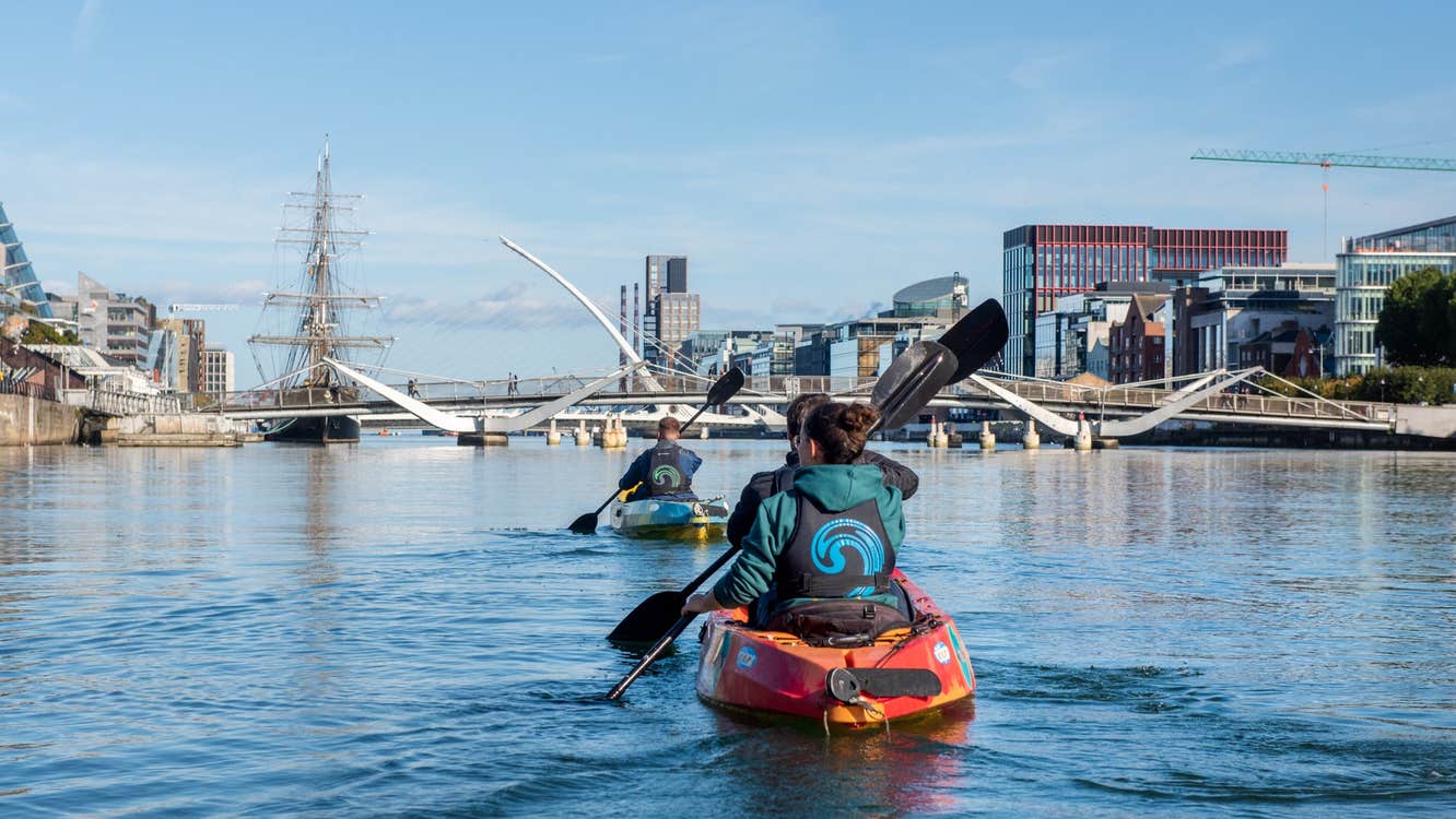 Two kayaks with three people paddle on the River Liffey towards the Samuel Beckett Bridge