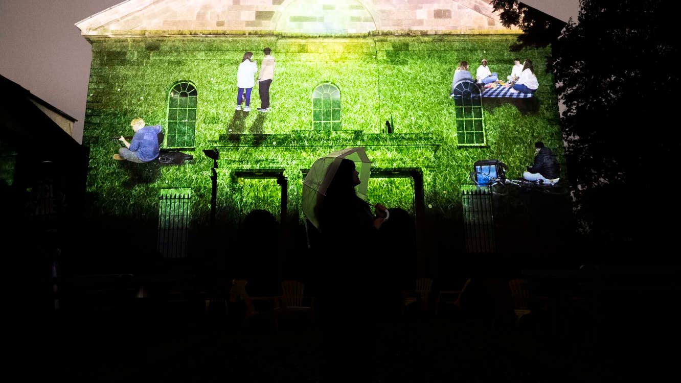 A projection of people on a field against the wall of a church. The silhouette of someone can be seen at the bottom of the image.