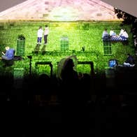 A projection of people on a field against the wall of a church. The silhouette of someone can be seen at the bottom of the image.