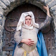 A heavily pregnant woman in medieval dress is standing in a stone archway.