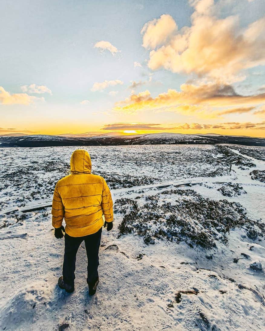 Person standing on snowy Ticknock with their back to the camera while the sunsets in the distance.