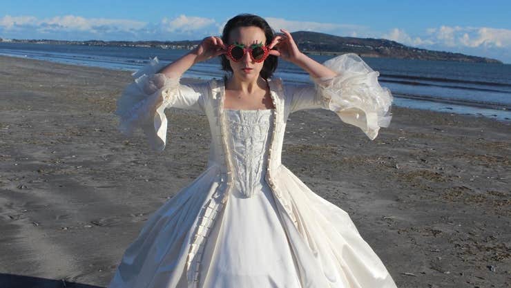 A woman in white, long 18th century dress holding on strawberry shaped sunglasses standing on a beach with the tide out.