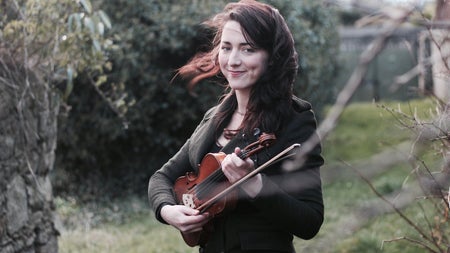 A woman standing beside a tree with a fiddle in her hand