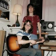 A standing and seated man with a guitar, beside a keyboard in a messy room