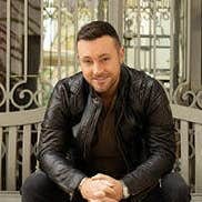 A smiling man dressed in black seated on a bench in front of metal railings