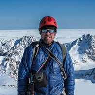 A man in outdoor clothing and mirrored sunglasses is standing against background of snowy mountains.