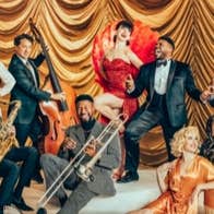 A group of musicians with instruments in vintage suits and dresses, some seated or standing or laying on a light coloured carpet.
