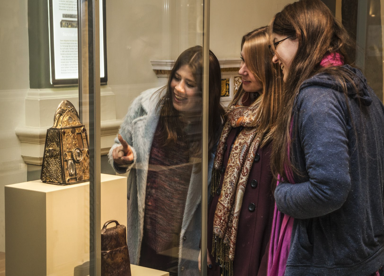 3 women are smiling, one pointing at a gold object in a glass display cabinet which they are gathered around.