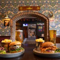 Two plates of burgers and chips with drinks on a bar counter framed by a patterned wallpaper and brick archway