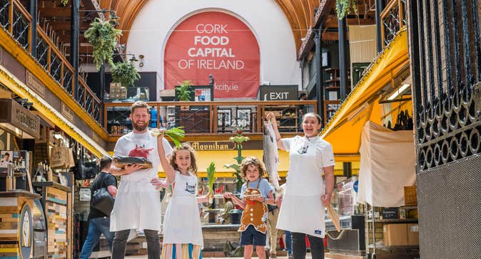 Group standing inside a market hall. One adult holds a tray of raw meat, a child holds green onions, another child holds baked goods, and one adult holds a large fish. A banner reading “CORK FOOD CAPITAL OF IRELAND” hangs above.