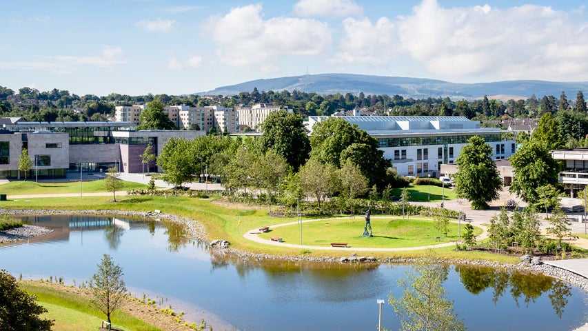 The grounds of University College Dublin with large buildings overlooking a pond and trees