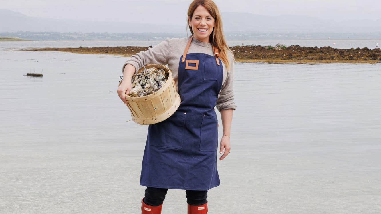Person standing in a grey sea holding a wooden bucket of oysters