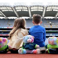 Rear view of 2 young children seated on the ground looking at the large stands in a stadium with multicoloured stuffed toys seated beside them.