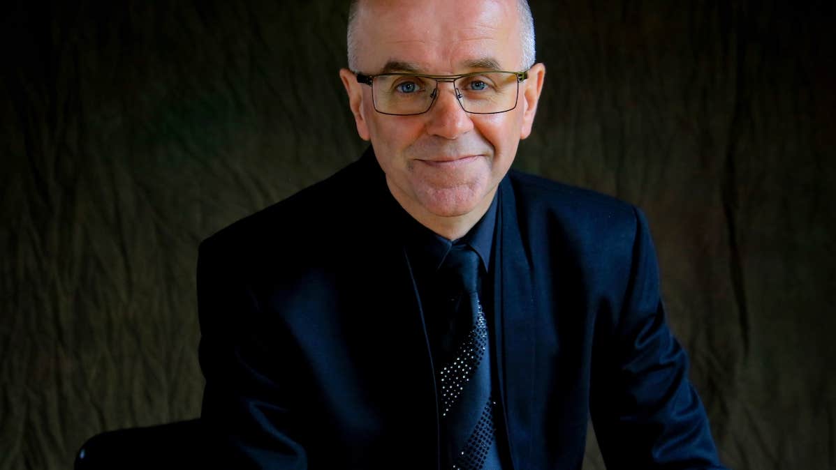 A smiling man in dark suit, tie and jacket against a dark, plain background.