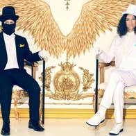 2 men seated in ornate gold chairs, one in black suit with face covered by a mask, the other in a white suit, against ornate white and old decorated wall.