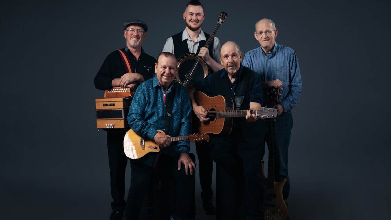 Group of 5 smiling musicians with their instruments in a tight group, against plain dark background.