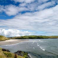 View of Inchydoney Strand with bright green grass, blue sky with some clouds, and the Atlantic ocean lapping at the sand.