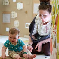 A smiling young baby seated on the floor looking down at something in front of them with an adult seated behind them.