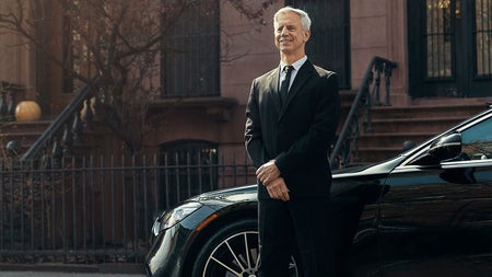 Person in a dark suit standing by a car on a street with steps in the background