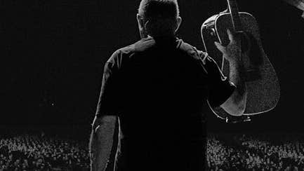 Darkly lit rear view of man holding up a guitar looking out over a large audience.