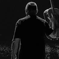 Darkly lit rear view of man holding up a guitar looking out over a large audience.