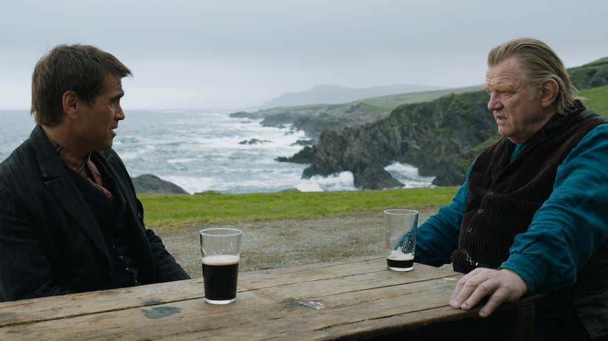 Brendan Gleeson and Collin Farrell from 'The Banshees of Inisherin' sit at a table with a scenic background, drinking pints of Guinness in a scene from the film.