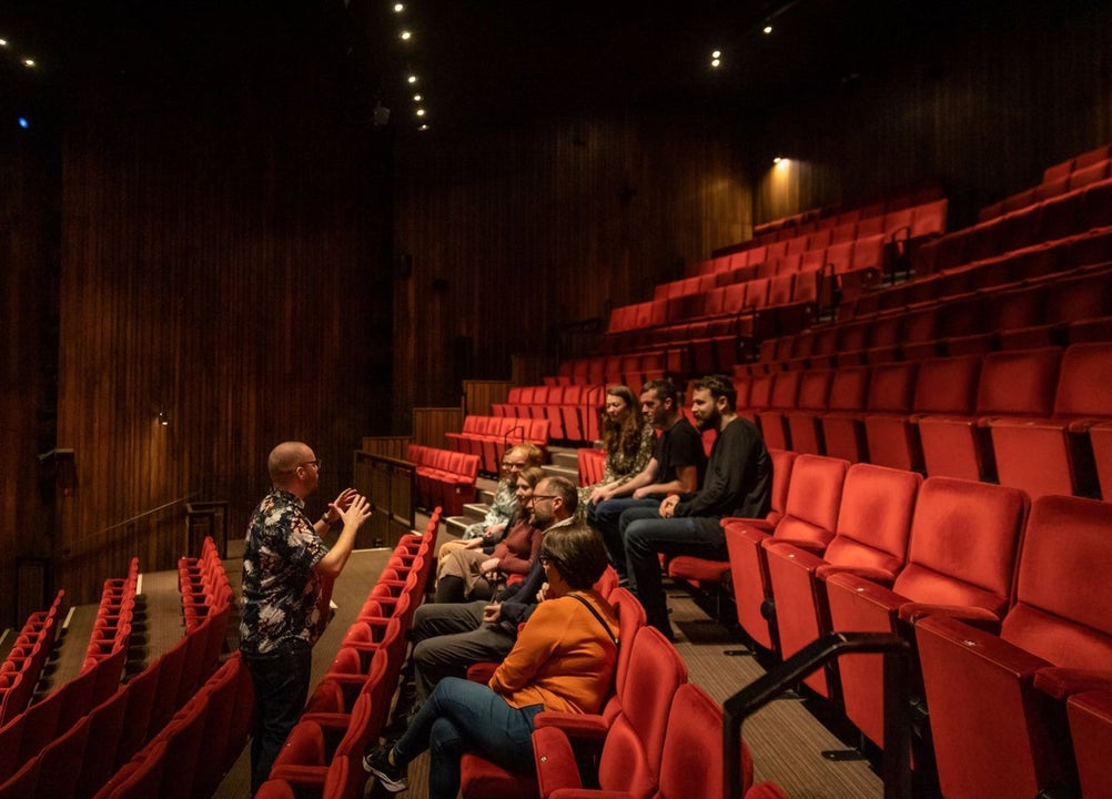 Small group of people sitting in a theatre auditorium with a tour guide