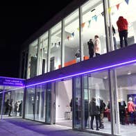 The exterior of the Swift Cultural Centre at night with people inside a well lit lobby and waiting area