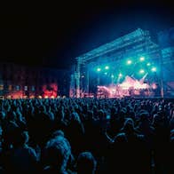 Distant view of large outdoor stage at night with large audience watching musicians.