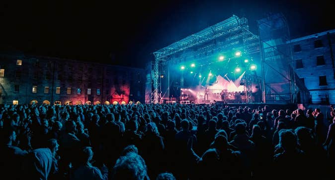 Distant view of large outdoor stage at night with large audience watching musicians.