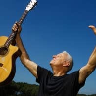 A man with arms outstretched, holding up a guitar in his right hand against blue sky