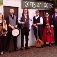 A line of 6 smiling men and women holding musical instruments standing in front of an old cottage