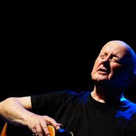 A bald man singing with eyes closed, holding a guitar.