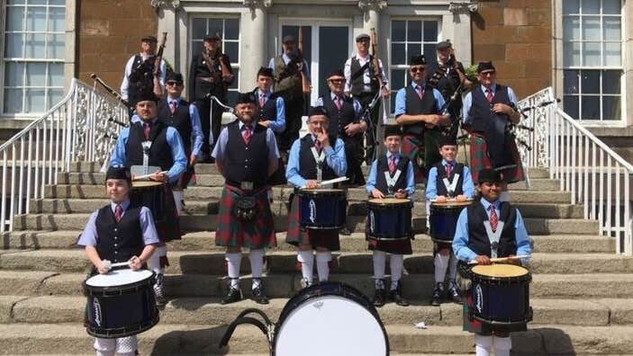A group of people with drums dressed in blue kilts standing scattered around a flight of stone steps in front of a building