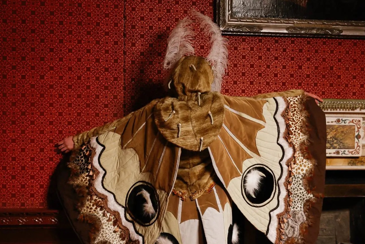 Reverse view of person with arms outstretched in ornate costume in shades of brown, white and cream to look like a moth.