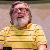 An Evening with Ricky Tomlinson, My Arse - a laughing older man with bushy beard seated with a mug on a table in front of him.