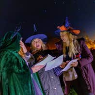 Adults in costume read their treasure hunt clues by torchlight at Nore Valley Park's Halloween Fire Trail, with warm lights glowing through the trees behind them.