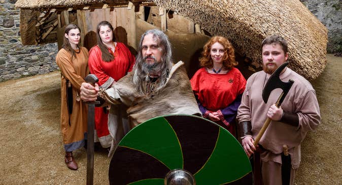A group of Viking reenactors stand in front of a building carrying a sword, shield and axe.