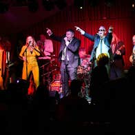 A group of 11 men and women singing and playing instruments with passion on a small stage.